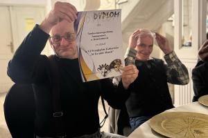 Dwóch panów siedzących za stołem pozuje do zdjęcia, jeden z nich trzyma dyplomm