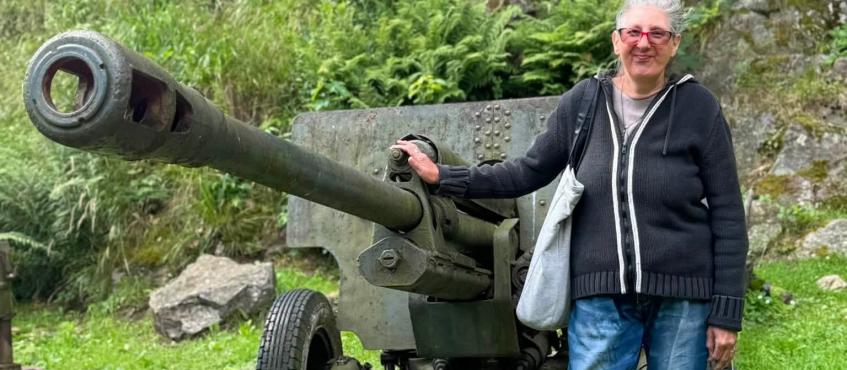 Na pierwszym planie, wśród zieleni pozuje kobieta trzymająca działo z długą lufą.